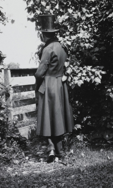 A man dressed in nineteenth century clothing, including a long trench-coat and hat, appears to stand in the garden facing away.