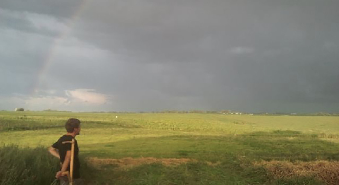 This is a picture of a field, with cloudy sky and rainbow. The image features a person standing in the front.