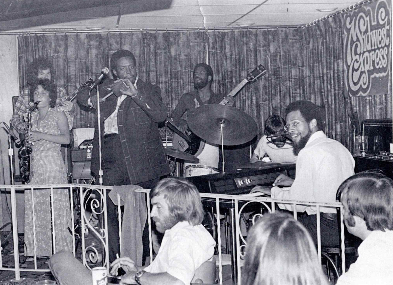  Midwest Express playing a gig in 1976. From far left, Marcia Miget is on saxophone, with Danny Nicholson behind her on guitar and Big Mike Edwards next to her at the mic. Dartanyan Brown (against the back wall) is on bass, John Grguric is on drums, and Bobby Parker is seated at the keyboard